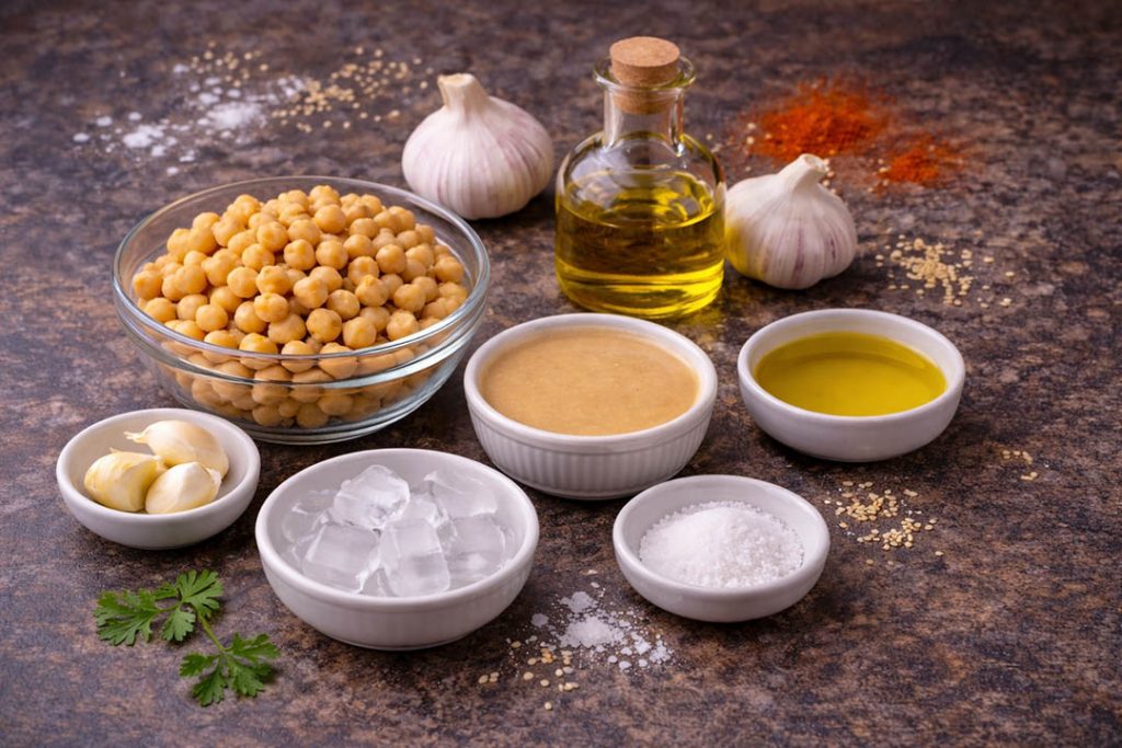 Ingredients for authentic Palestinian Lebanese hummus including chickpeas, tahini, garlic, olive oil, salt, and ice cubes arranged on a dark background.