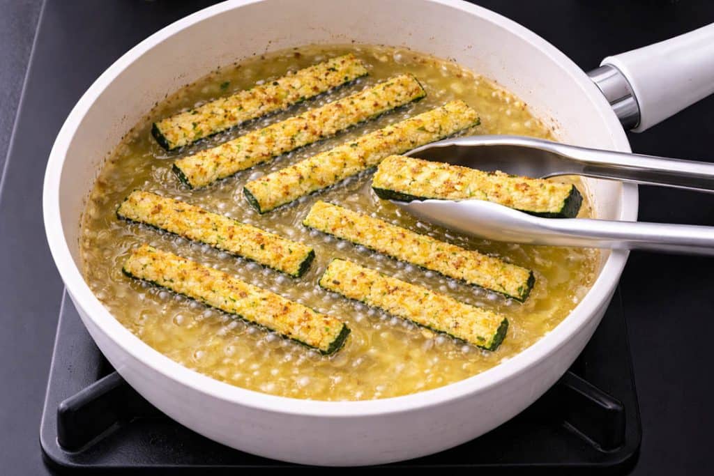 Parmesan garlic courgette zucchini sticks frying in oil in a white ceramic pan on a black hob