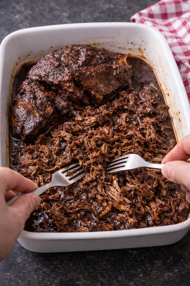 Slow-cooked smoky barbecue pulled beef being shredded with forks in a baking dish.