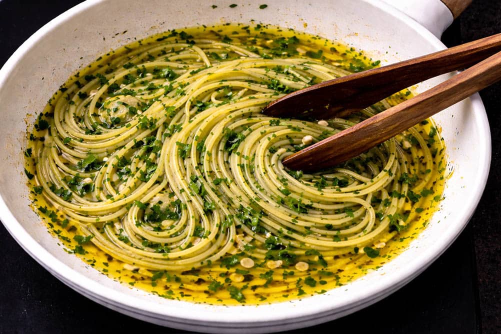 Spaghetti aglio e olio cooking in olive oil with chopped parsley in a white ceramic frying pan with wooden tongs, top view.