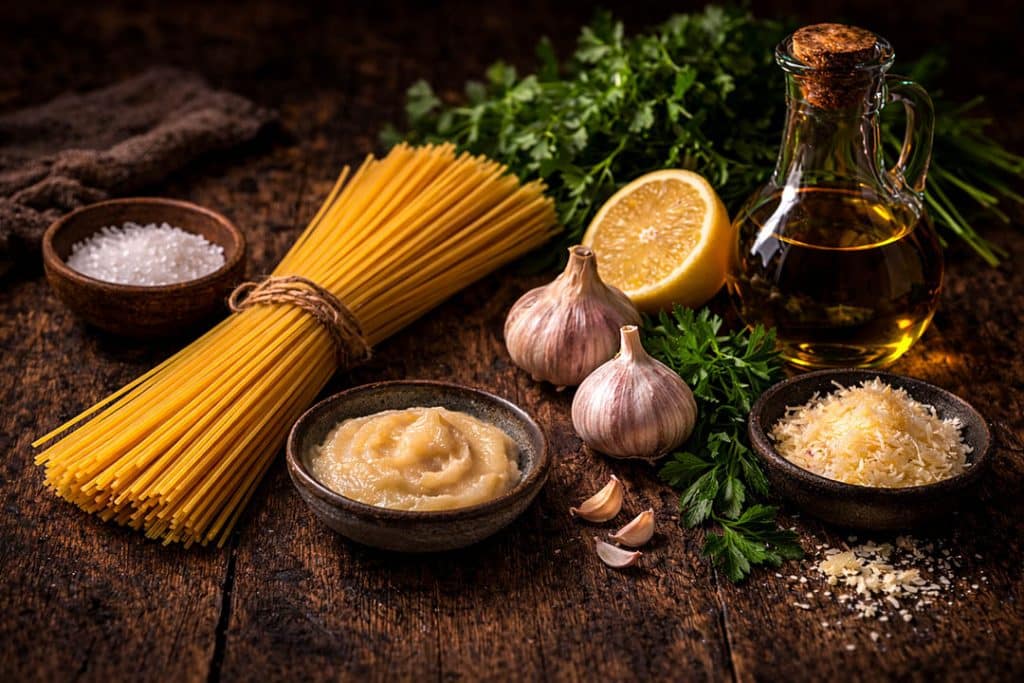 Spaghetti aglio e olio ingredients including spaghetti, garlic, parsley, lemon, Parmesan cheese, sea salt and extra virgin olive oil on a rustic wooden background.